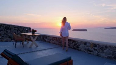 Huzurlu giyinmiş sakin bir kadın deniz manzaralı bir terasta güneşin batışından zevk alır.