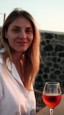 Beyaz gömlekli bir kadın gün batımında verandada şaraptan zevk alır, akşam ışığında huzur saçar.