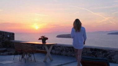 Beyaz gömlekli bir kadın deniz kenarında gün batımını seyrediyor elinde bir şarap kadehi, huzurlu ve rahatlatıcı..