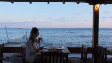 Gün batımında deniz kenarında bir restoranda yemek yiyen bir kadın. Arka planda sakin bir deniz manzarası var..