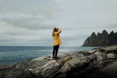 Parlak sarı bir ceket giyen bir kişi kayalık arazide duruyor, dağlarla birlikte denize bakıyor ve bir kamera kullanarak fotoğraf çekiyor. Lofoten, Norveç.
