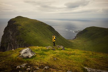 Dağ zirvesinde yalnız bir figür, sakin bir manzaradaki nefes kesici okyanusa düşünceli bir şekilde bakar. Lofoten, Norveç.