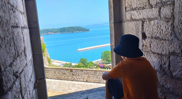 Çocuk denize bakan kalenin penceresinden dışarı bakar. Güzel deniz manzarası ve yeşil ağaçlarla kaplı küçük bir dağ.