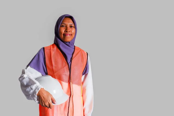 確信したイスラム教徒の女性労働者はカメラに微笑み 安全ヘルメットを持って来て 白い安全ヘルメット オレンジ色のベストとユニフォーム 隔離された灰色の背景 — ストック写真