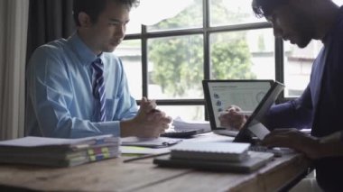 Takım elbiseli iki Asyalı iş adamı ofiste finans piyasası verilerini tartışıyor. İş adamları grafiklerde görülen istatistik dinamiklerini analiz ediyor.
