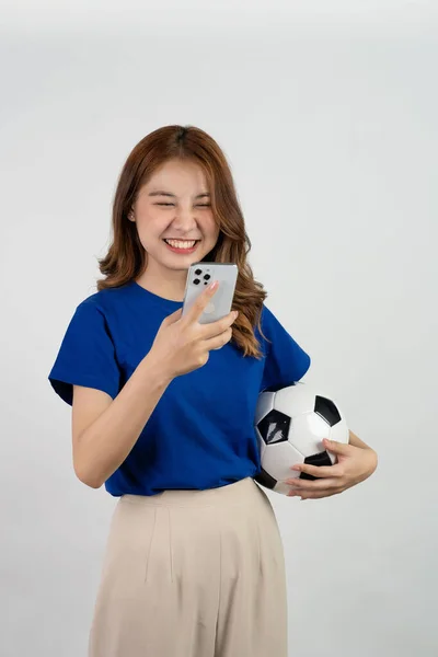 Happy Asian female soccer fan sending support to favorite team with soccer ball, smiling woman in blue t-shirt holding soccer ball to cheer at soccer game, isolated on white background.