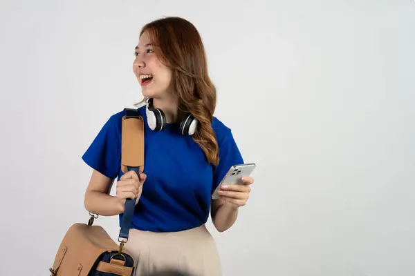 白い背景に隔離された立っている間青いシャツおよびバックパックでスマートフォンを使用するアジアの少女 — ストック写真