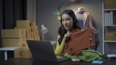 Asyalı kadın elbise stoklarını ve çevrimiçi perakende işini kontrol ediyor, İnternet satış sitesinde kıyafet satıyor, canlı yayınlar 4K video, moda tartışmaları ve çevrimiçi mağazada giysiler satıyor.