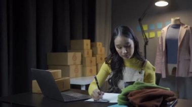 Asyalı kadın elbise stoklarını ve çevrimiçi perakende işini kontrol ediyor, İnternet satış sitesinde kıyafet satıyor, canlı yayınlar 4K video, moda tartışmaları ve çevrimiçi mağazada giysiler satıyor.