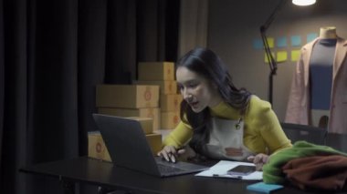 Asyalı kadın elbise stoklarını ve çevrimiçi perakende işini kontrol ediyor, İnternet satış sitesinde kıyafet satıyor, canlı yayınlar 4K video, moda tartışmaları ve çevrimiçi mağazada giysiler satıyor.
