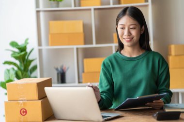 Arka planda paketlerle dolu bir evde oturan Asyalı genç bir kadının portresi. E-ticaret ve KOBİ 'ler için dizüstü bilgisayar ve hesap makinesi ile sipariş notları yaz..