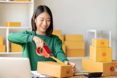Arka planda paketlerle dolu bir evde oturan Asyalı genç bir kadının portresi. E-ticaret ve KOBİ 'ler için dizüstü bilgisayar ve hesap makinesi ile sipariş notları yaz..