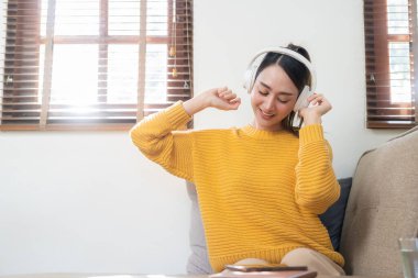 Modern Asyalı kadın, evdeki oturma odasındaki kanepede uzanıp müzik dinlerken kulaklık takıyor.