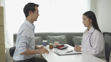 Beyaz üniformalı Asyalı kadın doktor iyi haberler veriyor. Sonuçları ya da belirtileri tartışıyor ve hastane sağlık kliniğindeki masada oturan erkek hastayla el ele tutuşuyor..