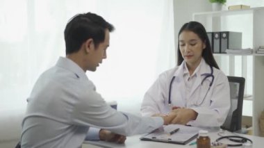 Beyaz üniformalı Asyalı kadın doktor iyi haberler veriyor. Sonuçları ya da belirtileri tartışıyor ve hastane sağlık kliniğindeki masada oturan erkek hastayla el ele tutuşuyor..