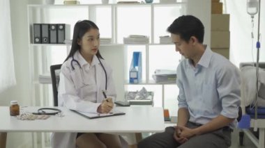 Beyaz üniformalı genç Asyalı doktor sağlık sorunlarını tartışmak için klinikteki erkek hastayla sonuçları ya da belirtileri tartışmak için pano kullanıyor..
