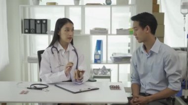Beyaz üniformalı genç Asyalı doktor sağlık sorunlarını tartışmak için klinikteki erkek hastayla sonuçları ya da belirtileri tartışmak için pano kullanıyor..