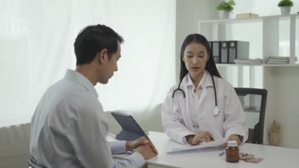 白い医学の制服の若いアジアの医師はクリップボードを使用して 健康問題を議論するためにクリップボードのテーブルに座っている男性患者と結果や症状を議論します — ストック動画