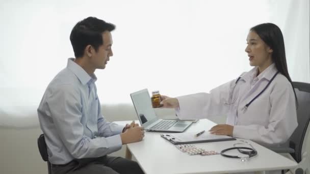 白い医学の制服の若いアジアの医師はクリップボードを使用して 健康問題を議論するためにクリップボードのテーブルに座っている男性患者と結果や症状を議論します — ストック動画