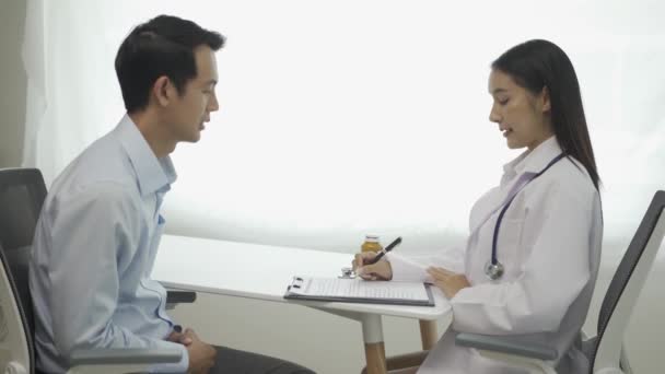 身穿白衣的年轻亚洲医生使用剪贴板与坐在诊所餐桌旁的男性病人讨论结果或症状 讨论健康问题 — 图库视频影像