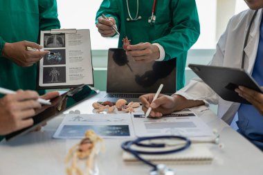 Çok ırklı bir tıbbi ekip beyaz laboratuvar önlüklü ve ameliyat önlüklü doktorlarla görüşüyor. Hasta kayıtlarını tartışmak ve bir tedavi bulmak için masalara oturuyorlar. Birlikte konuşan bir grup doktorun görüntüsü.