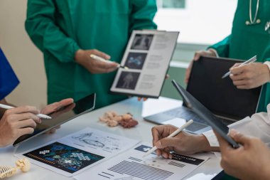 Çok ırklı bir tıbbi ekip beyaz laboratuvar önlüklü ve ameliyat önlüklü doktorlarla görüşüyor. Hasta kayıtlarını tartışmak ve bir tedavi bulmak için masalara oturuyorlar. Birlikte konuşan bir grup doktorun görüntüsü.