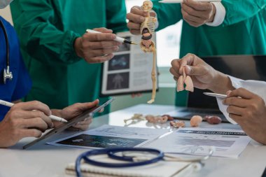 Çok ırklı bir tıbbi ekip beyaz laboratuvar önlüklü ve ameliyat önlüklü doktorlarla görüşüyor. Hasta kayıtlarını tartışmak ve bir tedavi bulmak için masalara oturuyorlar. Birlikte konuşan bir grup doktorun görüntüsü.