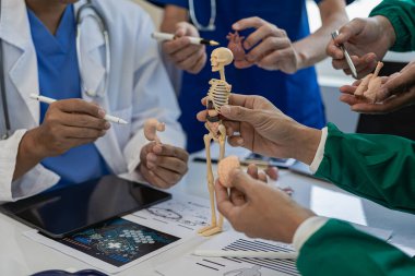 Çok ırklı bir tıbbi ekip beyaz laboratuvar önlüklü ve ameliyat önlüklü doktorlarla görüşüyor. Hasta kayıtlarını tartışmak ve bir tedavi bulmak için masalara oturuyorlar. Birlikte konuşan bir grup doktorun görüntüsü.