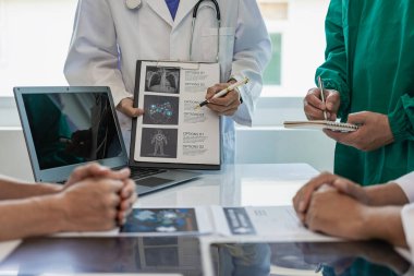 Çok ırklı bir tıbbi ekip beyaz laboratuvar önlüklü ve ameliyat önlüklü doktorlarla görüşüyor. Hasta kayıtlarını tartışmak ve bir tedavi bulmak için masalara oturuyorlar. Birlikte konuşan bir grup doktorun görüntüsü.