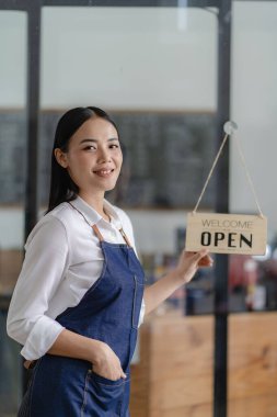 Güzel bir Asyalı işletme sahibi kapıda durup müşterilerden emir almaya hazırlanıyor. Önlüklü genç bir barista tablet tutuyor ve açık bir tabelayla kafenin kapısının önünde duruyor..