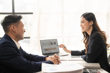 İki Asyalı iş adamı dizüstü bilgisayarlarla ve finansal belgelerle mutlu bir şekilde gülümsüyor. Ofiste projelerinden ve yaratıcı fikirlerden bahsediyorlar..