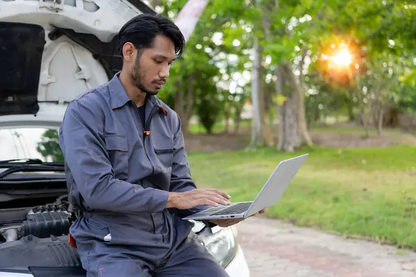 Auto mechanic inspects car by inspecting and writing checklist on clipboard. Mechanic checks car parts stock on laptop computer with car broken down on the side of the road