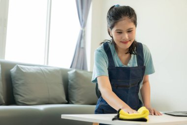 Temizlik üniformalı Asyalı bir kadın evi temizler ve oturma odasındaki mikrofiber bezle masayı siler ve temiz ve düzenli bir ev için yerleri süpürgeyle paspaslar..