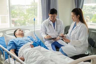 Asyalı erkek hasta, tıbbi sağlık konseptinin yanında danışmanlık ve yardım sağlayan doktor ekibiyle yatakta yatıyor..
