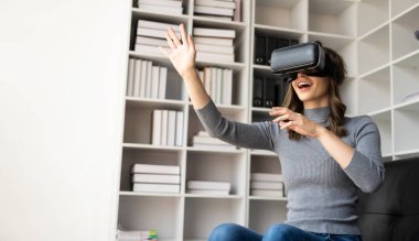 VR kulaklığıyla şaşırmış, havayı hisseden mutlu genç bir kadın. Gözlüklü gülümseyen beyaz kadın, sanal gerçeklik teknolojisi aygıtı, evdeki kanepede otururken 3D simülasyon videosu izliyor..