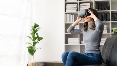 VR kulaklığıyla şaşırmış, havayı hisseden mutlu genç bir kadın. Gözlüklü gülümseyen beyaz kadın, sanal gerçeklik teknolojisi aygıtı, evdeki kanepede otururken 3D simülasyon videosu izliyor..