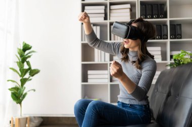 VR kulaklığıyla şaşırmış, havayı hisseden mutlu genç bir kadın. Gözlüklü gülümseyen beyaz kadın, sanal gerçeklik teknolojisi aygıtı, evdeki kanepede otururken 3D simülasyon videosu izliyor..