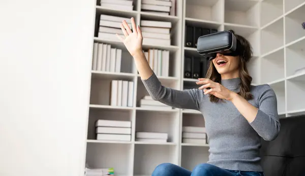 VR kulaklığıyla şaşırmış, havayı hisseden mutlu genç bir kadın. Gözlüklü gülümseyen beyaz kadın, sanal gerçeklik teknolojisi aygıtı, evdeki kanepede otururken 3D simülasyon videosu izliyor..