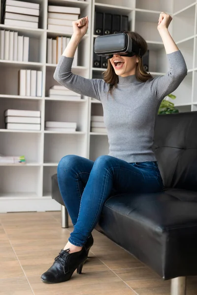 Happy young woman surprised in VR headset, feeling air. Smiling caucasian woman wearing vr glasses, virtual reality technology device, watching 3d simulation video while sitting on sofa at home.