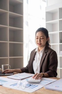 Güzel Asyalı iş kadını hesap makinesini kullanarak hesap tablosunu analiz ediyor ve finansal raporları analiz etmek için masa başında belgeler hazırlıyor.