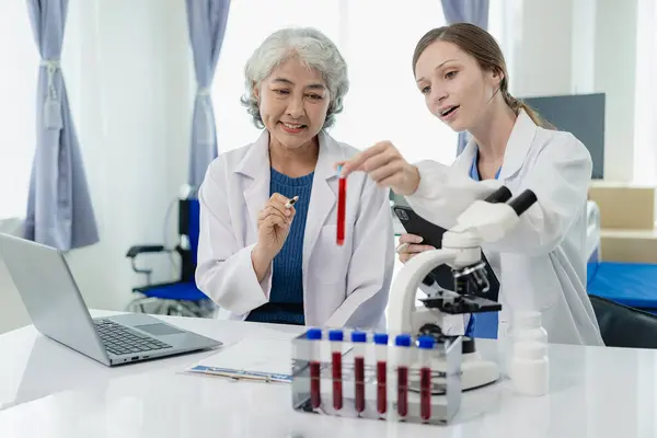 Laboratory, old female doctor and woman doing medical research with blood in tube, beautiful young woman in white coat sitting in office and working with blood sample in laboratory medical laboratory