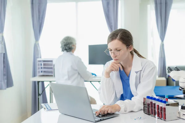 Laboratory, old female doctor and woman doing medical research with blood in tube, beautiful young woman in white coat sitting in office and working with blood sample in laboratory medical laboratory