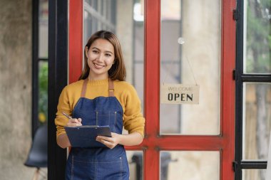 Genç bir Asyalı bayan girişimci yiyecek ve içecek dükkanının önüne bir hoş geldin tabelası astı. Güzel bir garson elinde bir tablet tutuyor ve restoranda yemek siparişi vermek için talimatlar hazırlıyor..