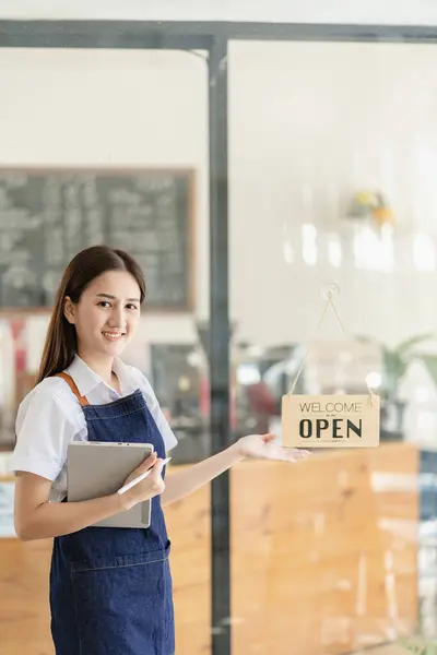 A young Asian female entrepreneur hangs a welcome sign in front of a food and beverage shop. A beautiful waitress holds a tablet and prepares instructions for ordering food in the restaurant.