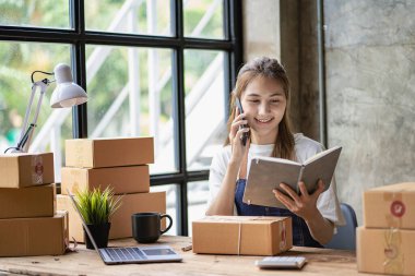 Küçük bir işletme kurun. Genç Asyalı kadın ev ofisinin bilgisayarları ve kutularıyla çalışan bir müşteriyle telefonda konuşuyor. Çevrimiçi pazarlama ve ambalaj kutusu lojistik kavramı KOBİ.