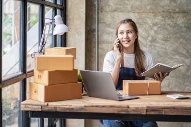 Küçük bir işletme kurun. Genç Asyalı kadın ev ofisinin bilgisayarları ve kutularıyla çalışan bir müşteriyle telefonda konuşuyor. Çevrimiçi pazarlama ve ambalaj kutusu lojistik kavramı KOBİ.