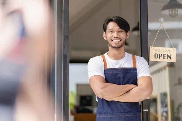 small business idea A smiling African American bakery worker in an apron stands outside a coffee shop with an open sign. Owner of a small coffee shop