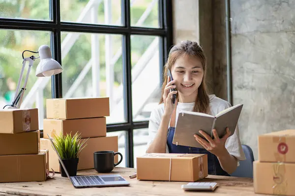 Küçük bir işletme kurun. Genç Asyalı kadın ev ofisinin bilgisayarları ve kutularıyla çalışan bir müşteriyle telefonda konuşuyor. Çevrimiçi pazarlama ve ambalaj kutusu lojistik kavramı KOBİ.