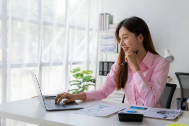 Genç Asyalı kadın masamda oturmuş elinde veri ve grafik kağıdıyla dizüstü bilgisayarını kullanıyor, hesapları analiz ediyor, iş geliştirme stratejileri planlıyor..