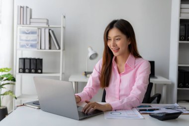 Genç Asyalı kadın masamda oturmuş elinde veri ve grafik kağıdıyla dizüstü bilgisayarını kullanıyor, hesapları analiz ediyor, iş geliştirme stratejileri planlıyor..
